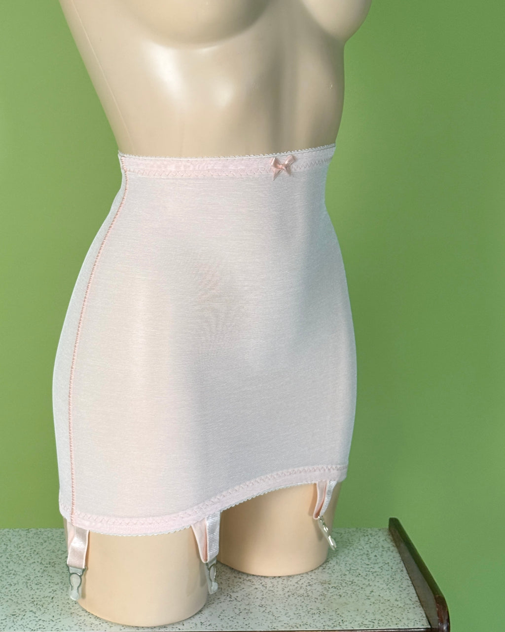 A light pink shapewear brief or girdle displayed on a beige mannequin torso against a solid green background.