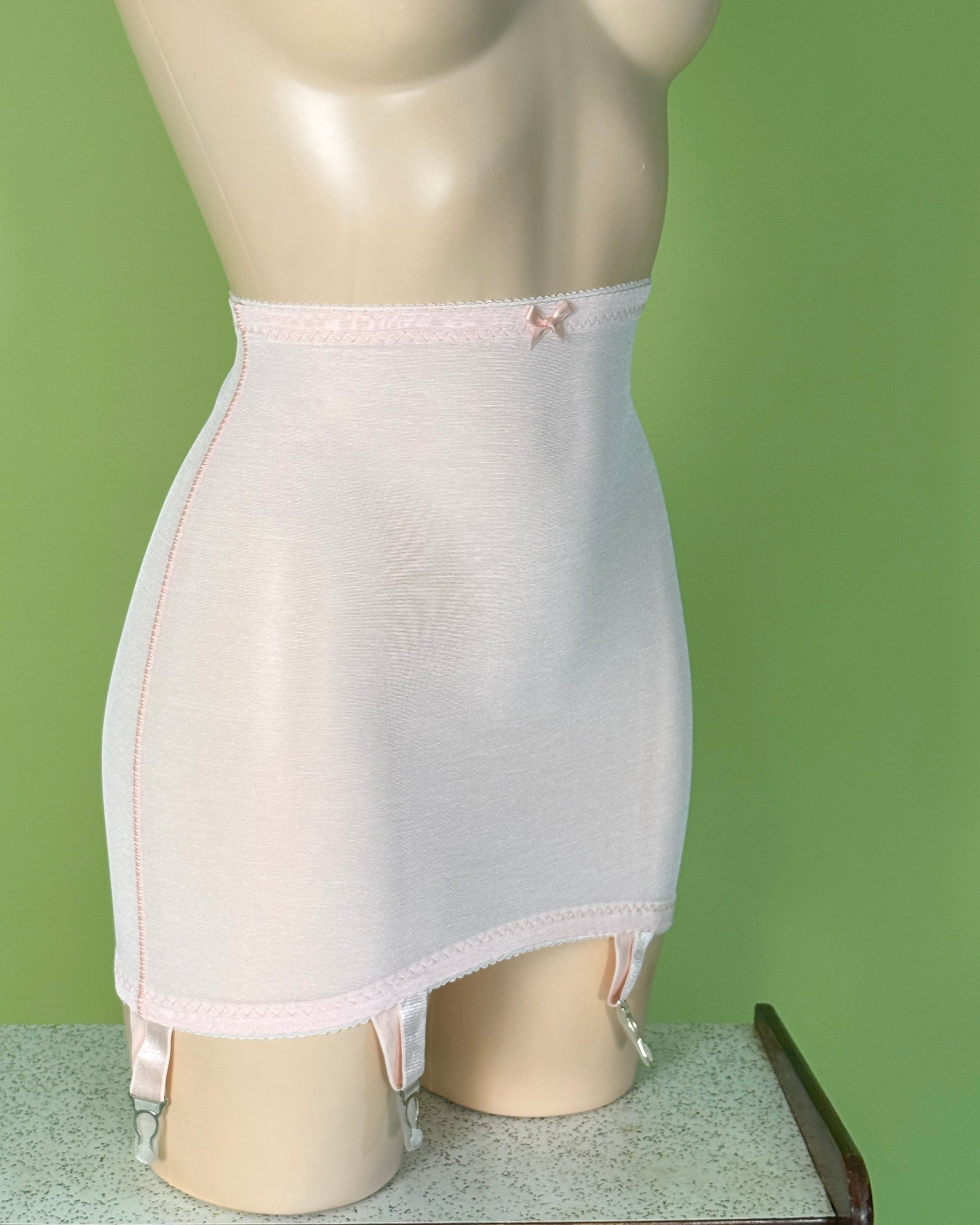 A light pink shapewear brief or girdle displayed on a beige mannequin torso against a solid green background.
