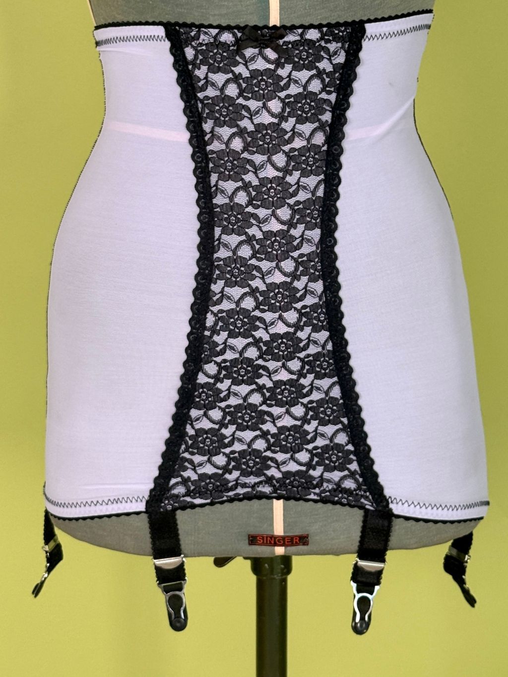Light purple girdle  with a central black lace panel and black garter straps, displayed on a gray dress form against a yellow-green background.