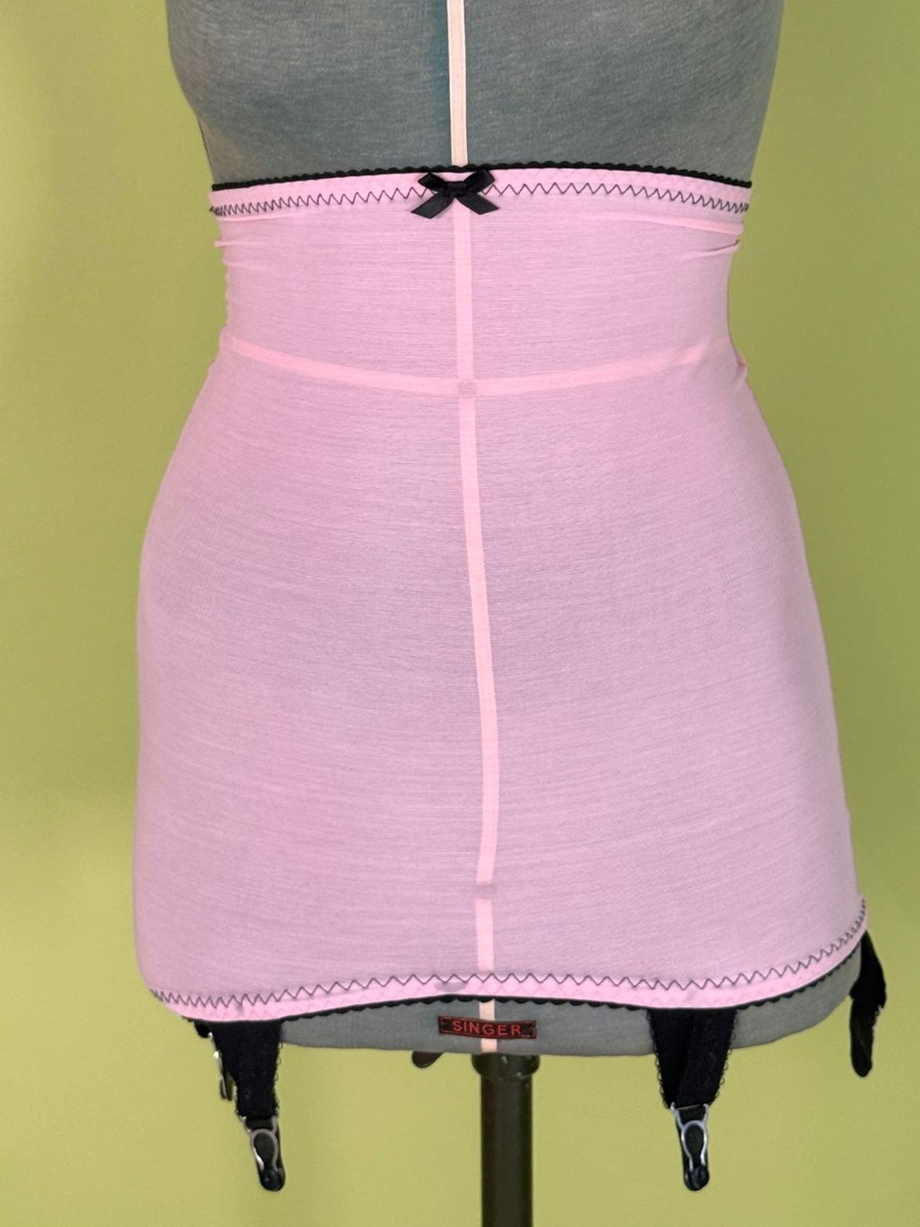 A light pink high waist girdle  displayed on a gray dress form mannequin against a solid olive green background.