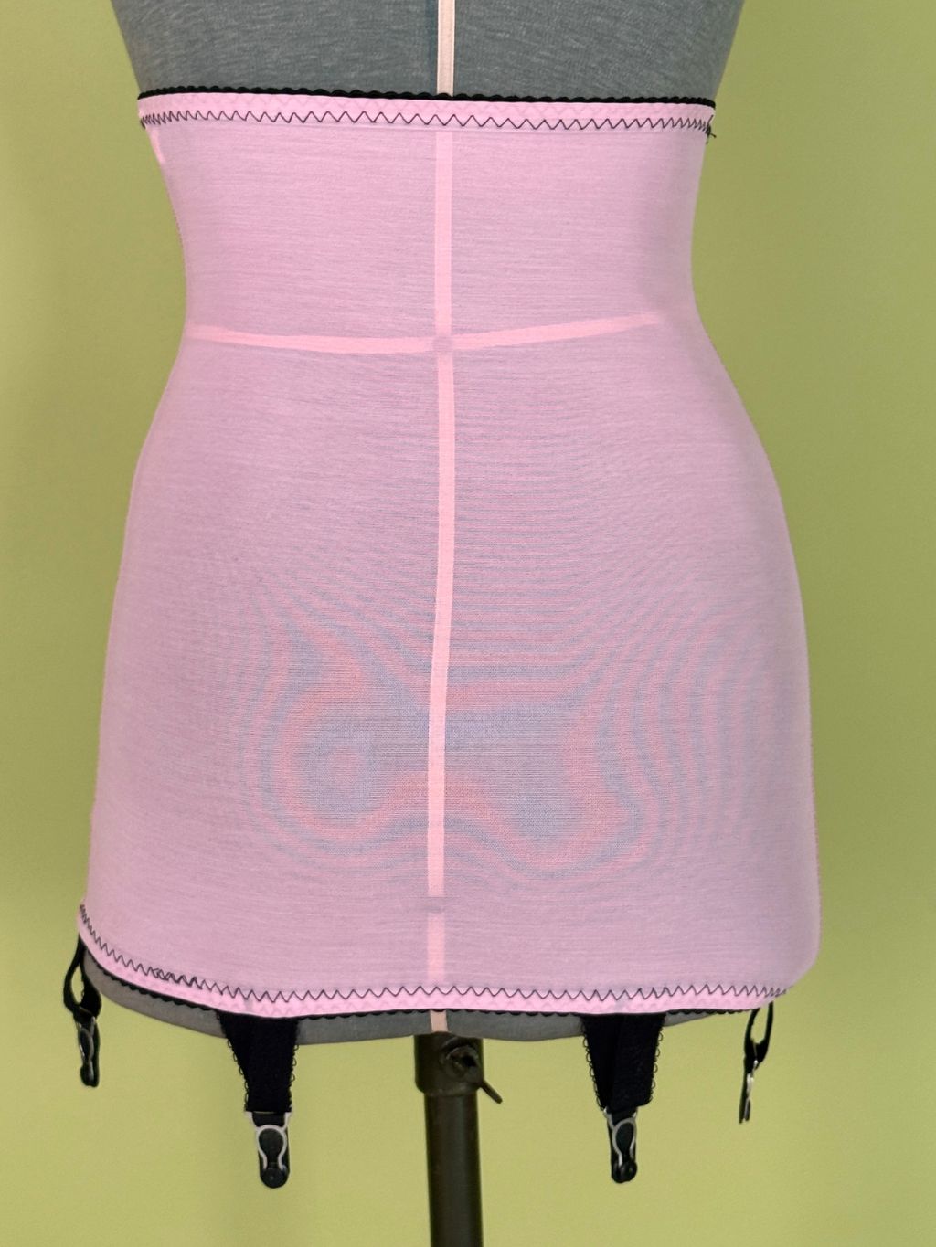 A pink high waist girdle  displayed on a gray dress form mannequin against a solid olive green background.