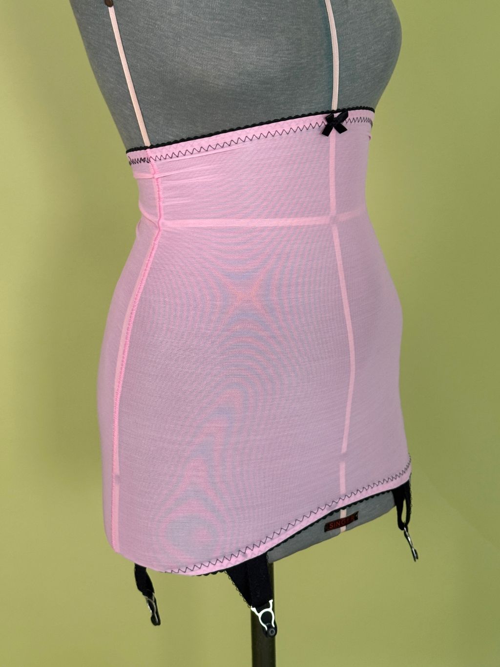 A light pink high waist girdle displayed on a gray dress form mannequin against a solid olive green background.
