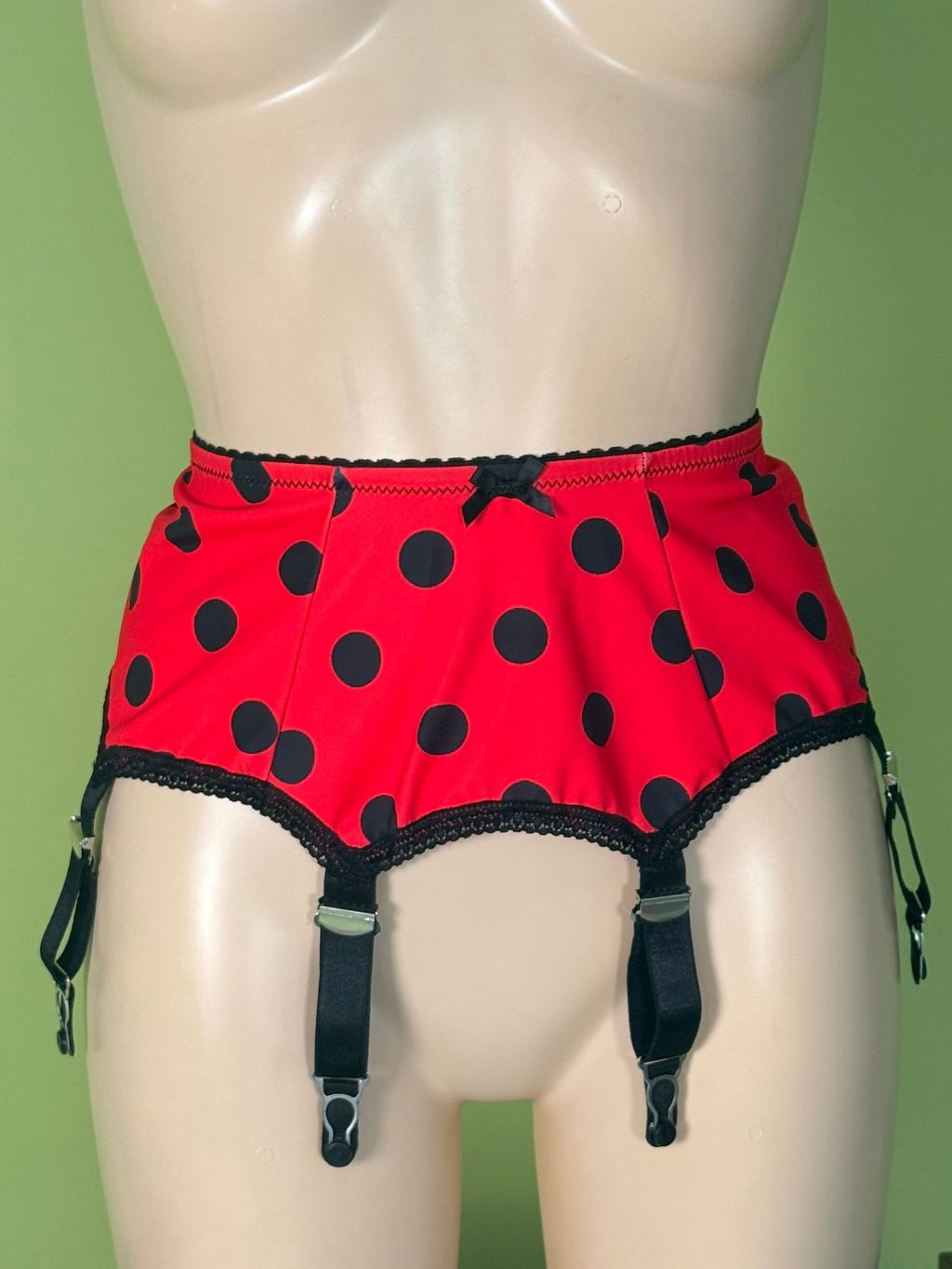 Red and black polka dot garter belt briefs displayed on a beige mannequin torso against a solid green background.