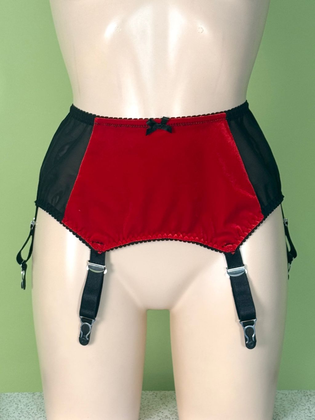 Red and black high-waisted garter belt with suspender straps, displayed on a beige mannequin torso against a light green background.