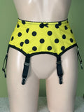 Yellow and black polka dot garter belt with black garter straps on a beige mannequin torso against a green wall.