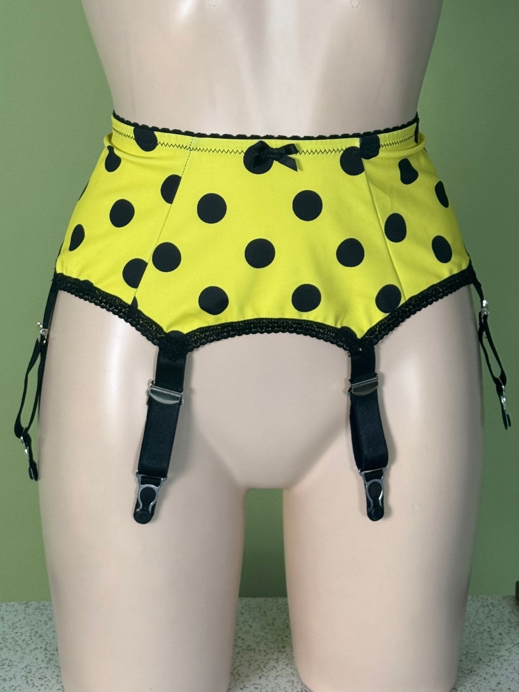 Yellow and black polka dot garter belt with black garter straps on a beige mannequin torso against a green wall.