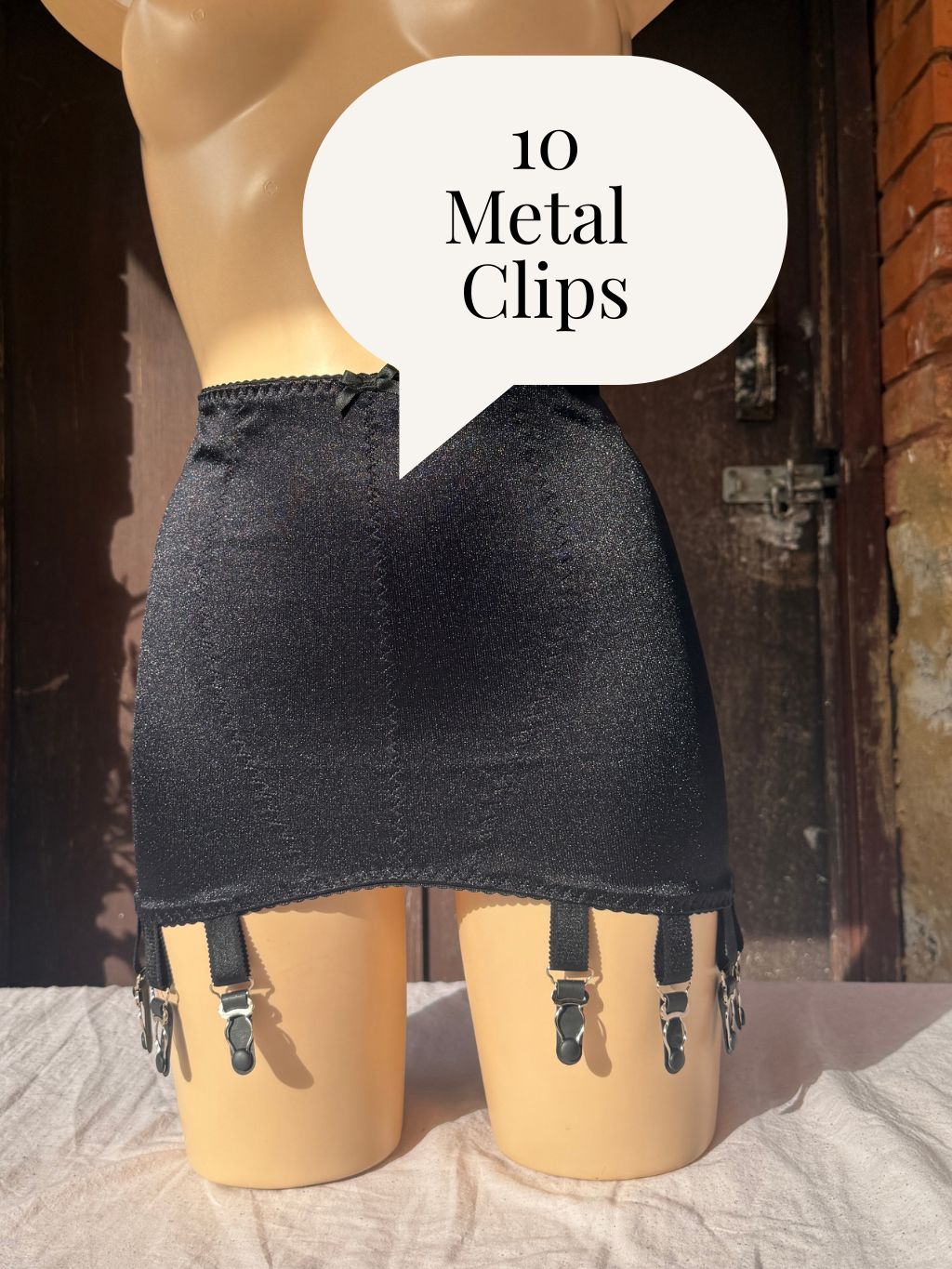 A black girdle with attached black garter straps and silver-toned metal clips, displayed on a beige mannequin. A white speech bubble overlay reads '10 Metal Clips'.