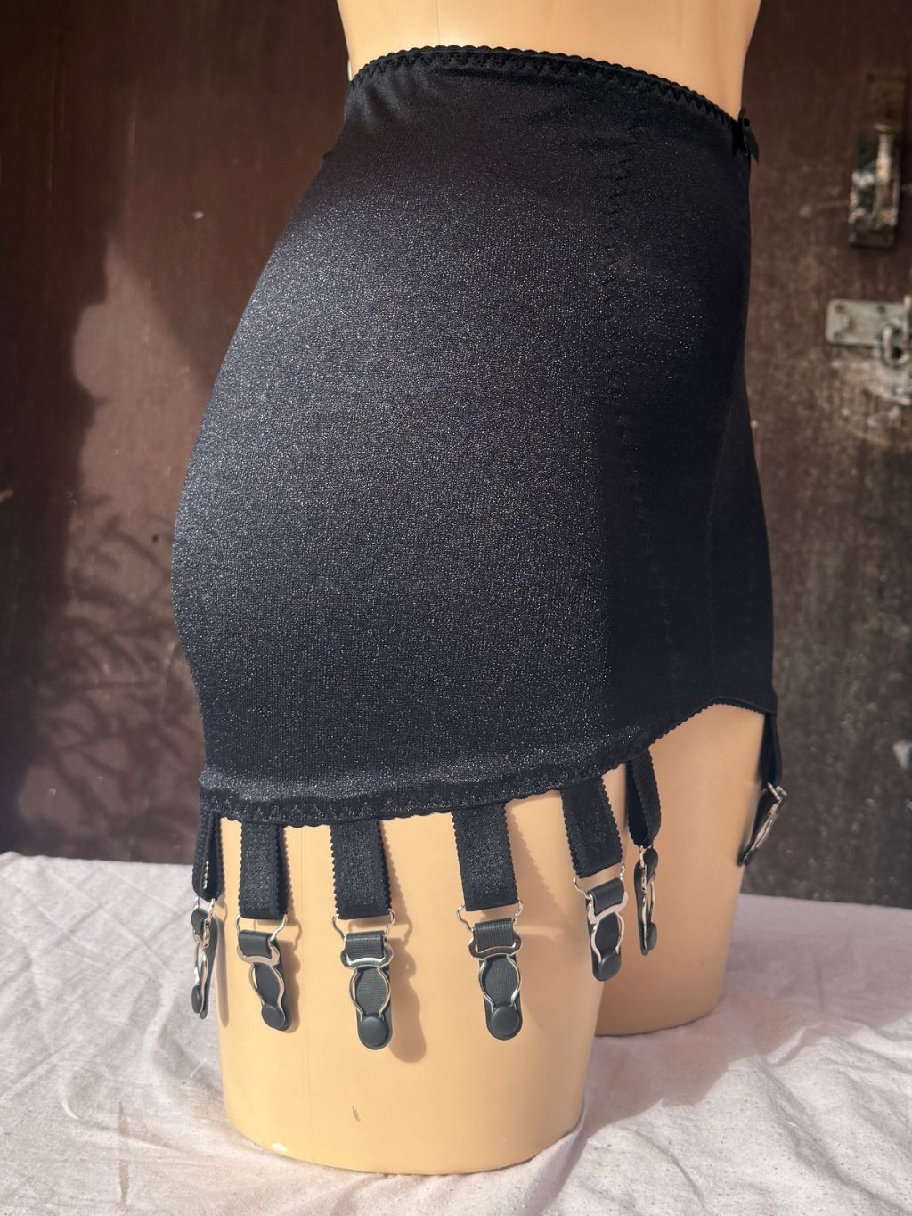 Black girdle skirt with multiple adjustable suspender straps and metal clips, displayed on a beige mannequin torso against a dark rustic background.