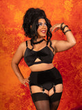 A woman modeling a black lingerie set consisting of a bralette with a front cutout, matching high-waisted briefs, and attached garter straps with thigh-high stockings, posed against a textured orange and red background.