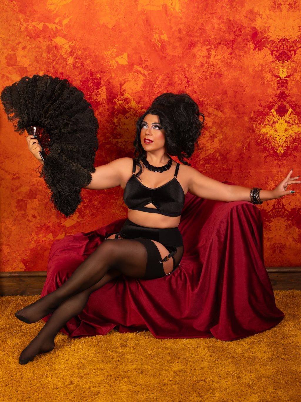 A person posed in a dramatic black lingerie set with sheer black tights, seated on a draped burgundy fabric prop against a textured orange wall, while holding a large black feather fan.