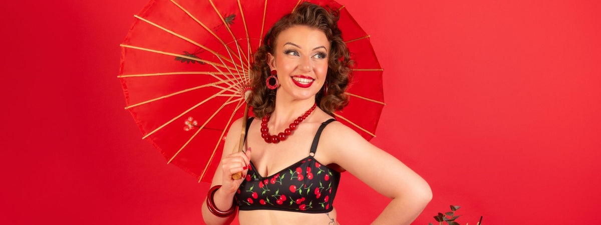 A woman with curly brown hair poses against a solid red background, holding a red Japanese-style paper umbrella. She is wearing a black floral bralette, a red beaded necklace, and red hoop earrings.