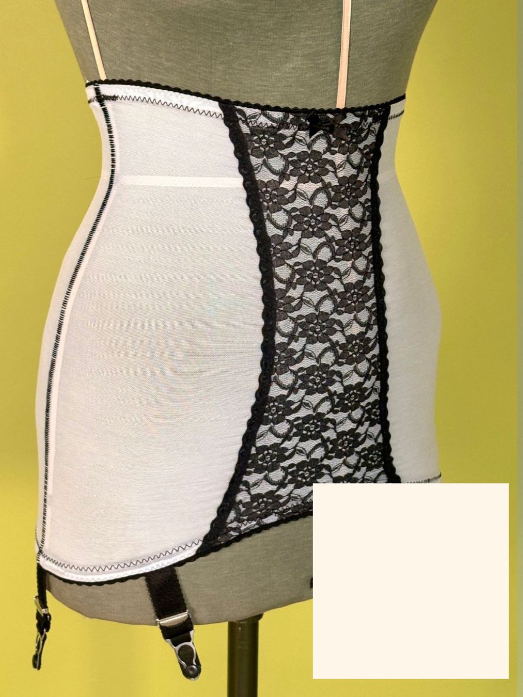 Cream and black girdle with garter straps, displayed on a gray dress form against a yellow-green background, with a color swatch inset in the bottom right corner.