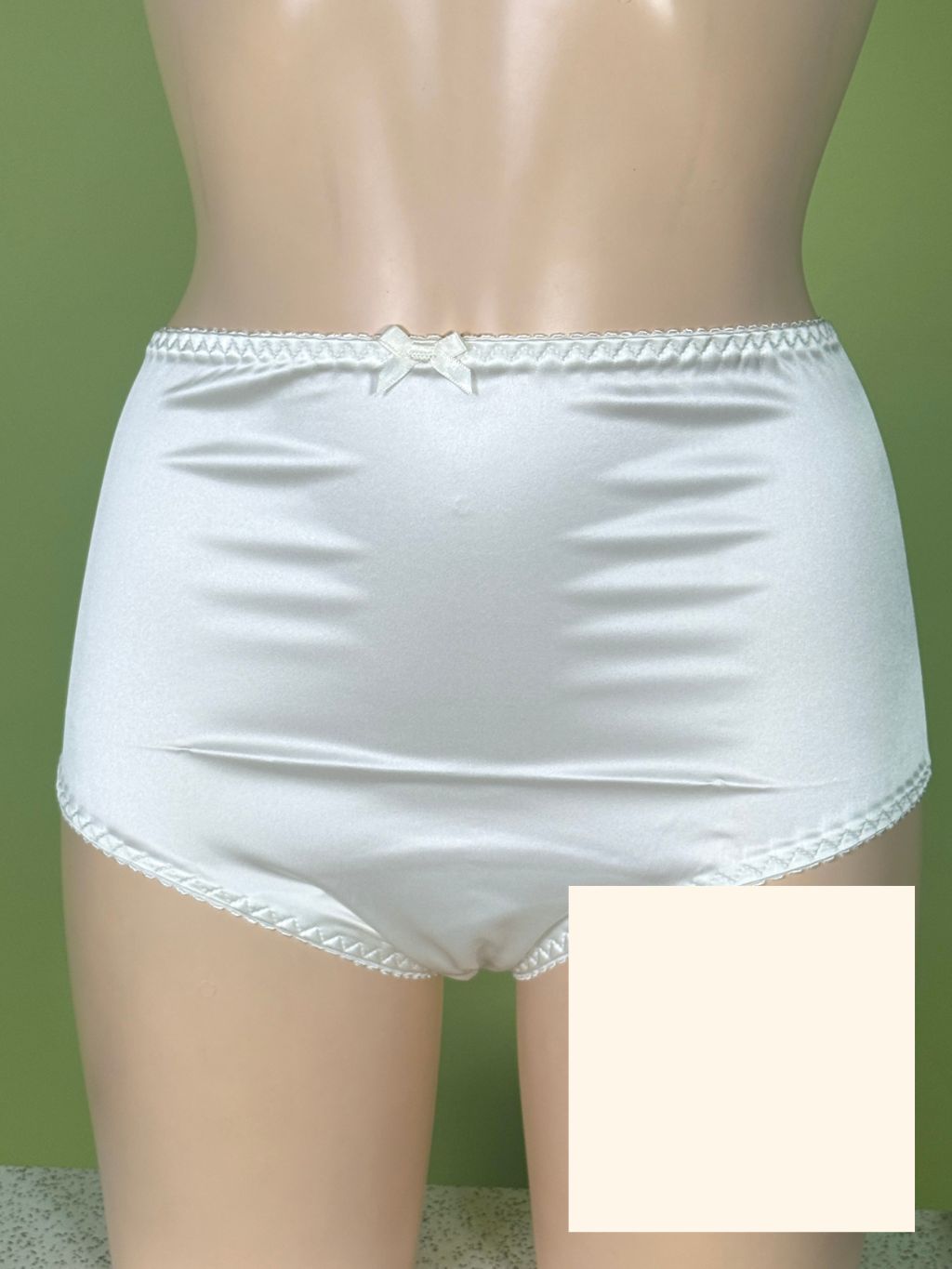 Cream satin briefs with lace trim and a small bow at the back waistband, displayed on a mannequin against a green background. A color swatch box is overlaid in the bottom right corner.