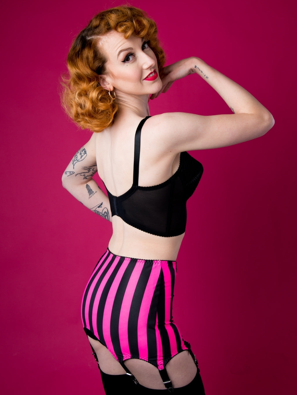 Woman wearing black top and black and pink striped girdle with black detailing on a red background.