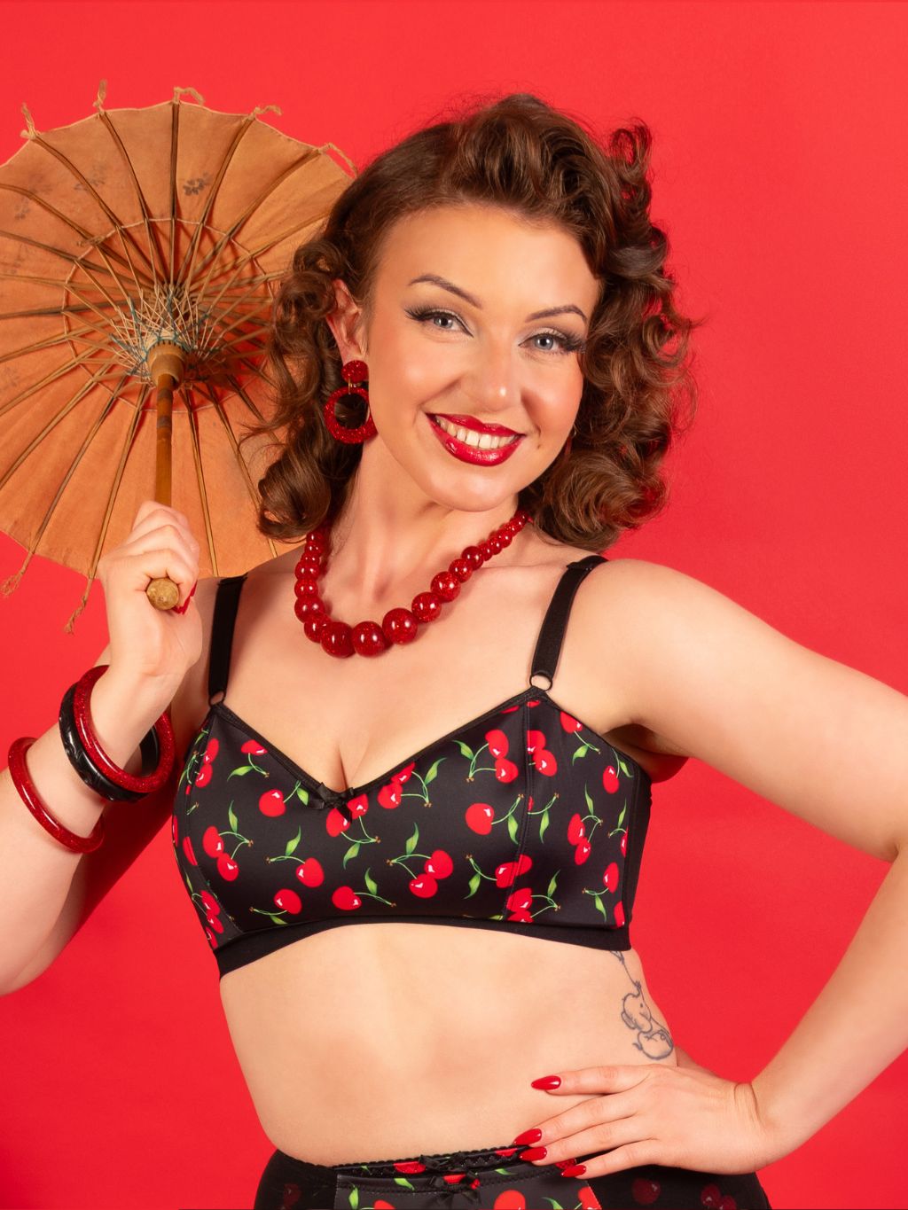 Woman in a black bralette with red cherry pattern holding a brown umbrella against a red background