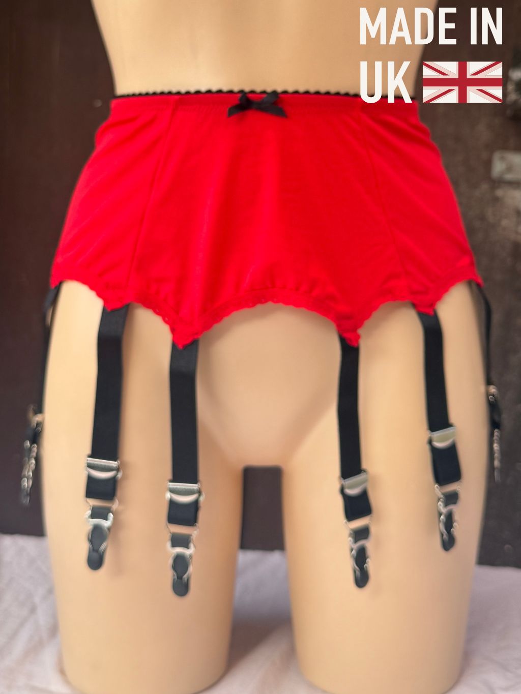 Red garter belt with black suspender straps and silver metal clips, displayed on a beige mannequin. A 'MADE IN UK' graphic overlay is in the top right corner.