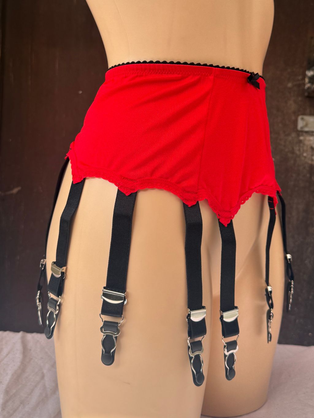 Red garter belt with black suspenders and silver metal clips, displayed on a beige mannequin torso against a dark brown background.