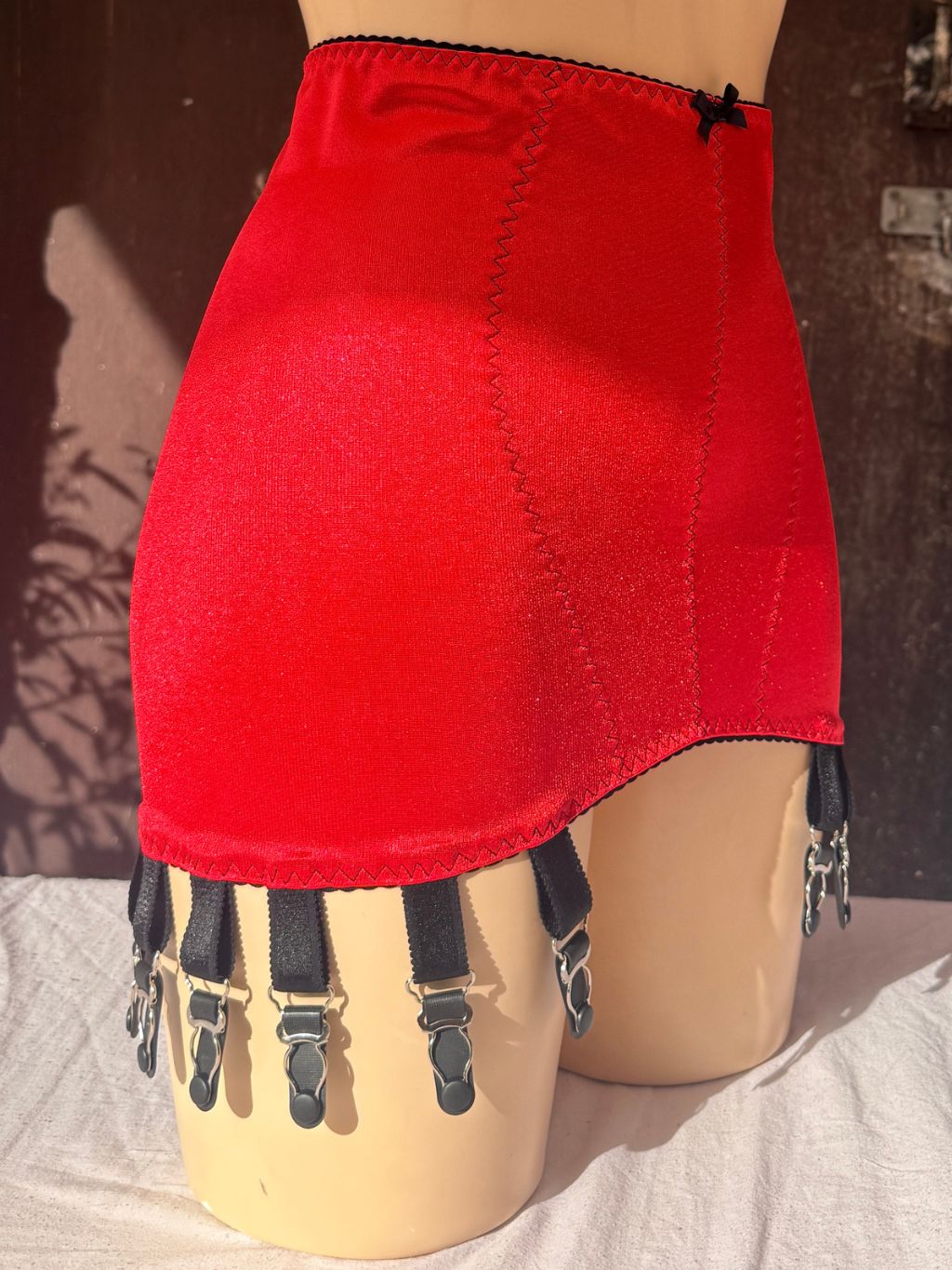 A bright red girdle displayed on a beige mannequin, featuring black suspender straps with silver metal clips and a small black bow at the waistband.