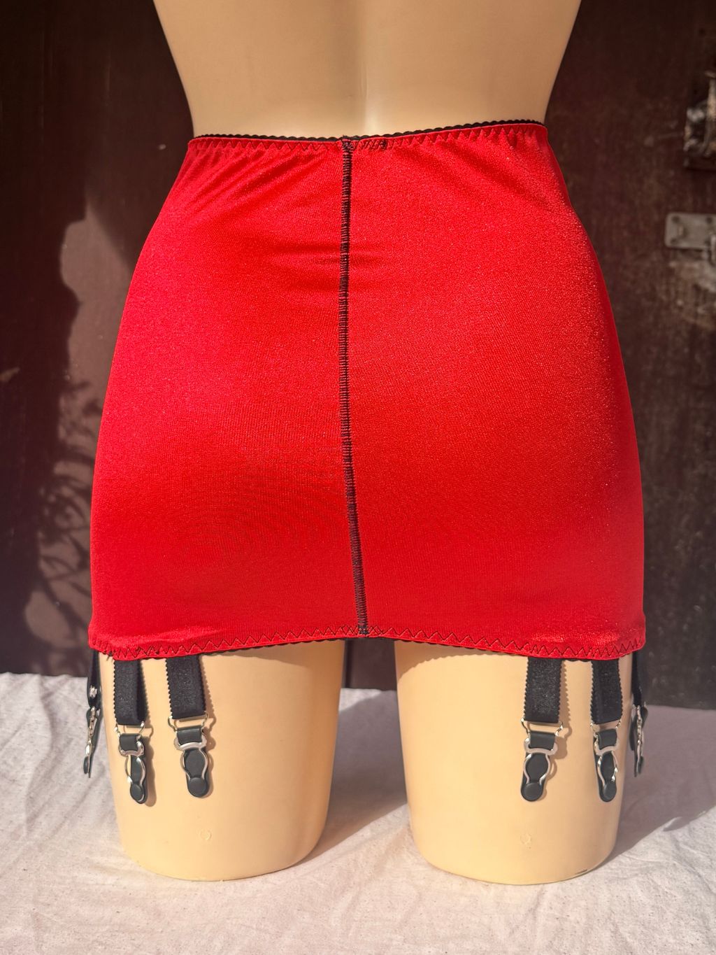 A bright red girdle displayed on a beige mannequin, featuring a center seam and attached black garter straps with metal clips at the upper thighs.