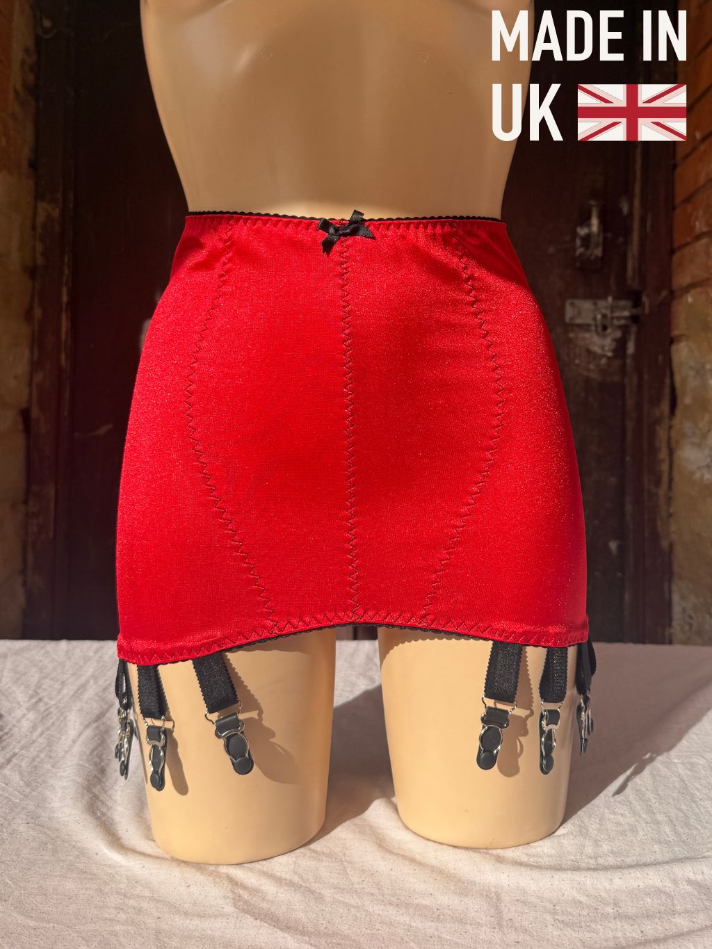 A red girdle with black suspender straps and clips, displayed on a beige mannequin. The background is a rustic dark wood door, and a 'MADE IN UK' graphic overlay is in the top right corner.