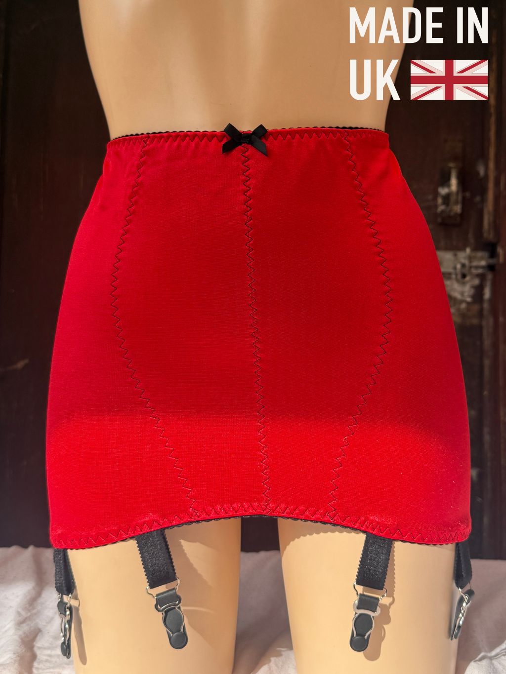 Red girdle with black garter straps and a small black bow at the center waistband, displayed on a mannequin with a 'MADE IN UK' badge in the top right corner.