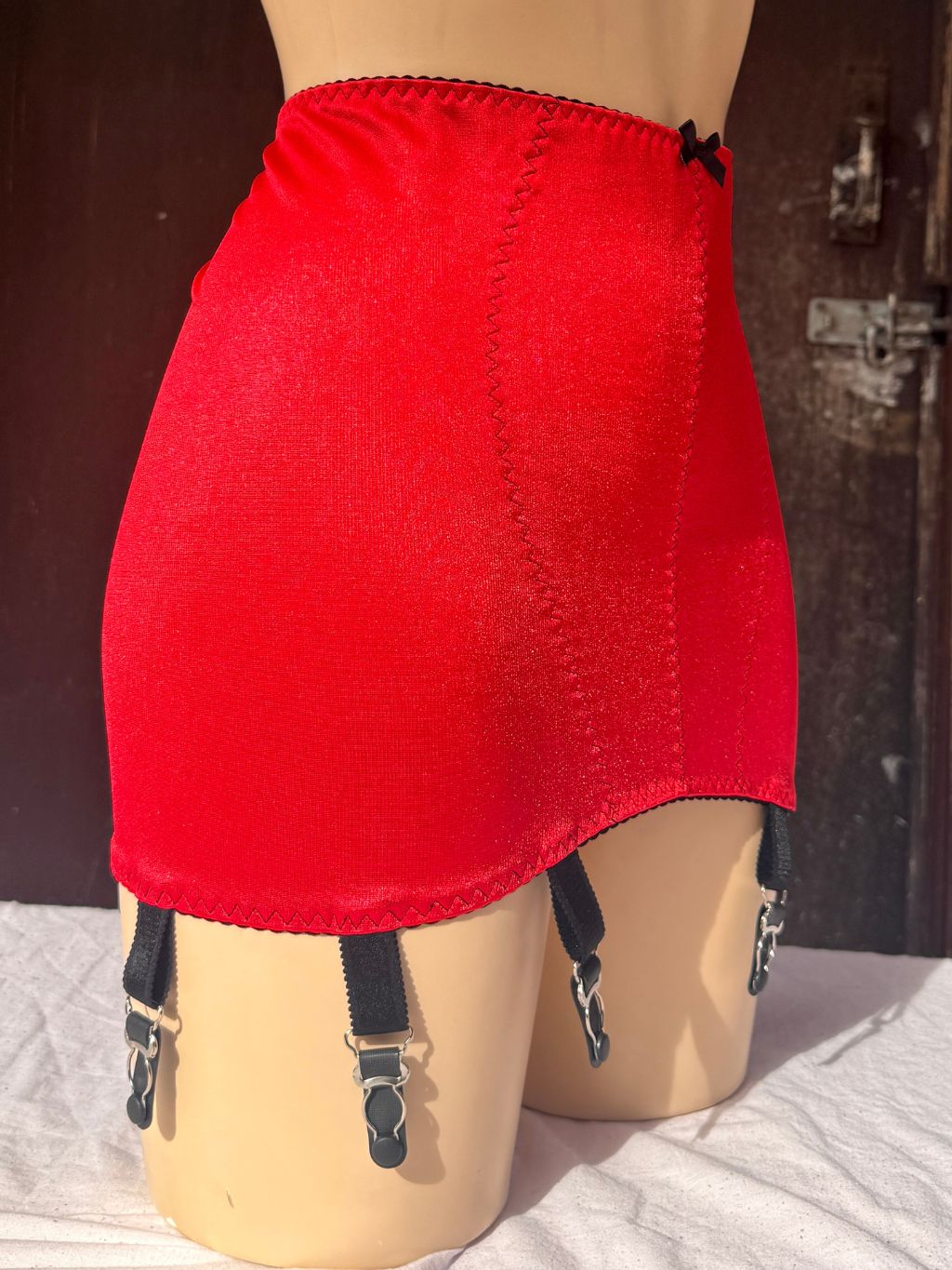 A bright red girdle with black suspender straps and silver metal clips, displayed on a beige mannequin leg against a dark wooden door background.