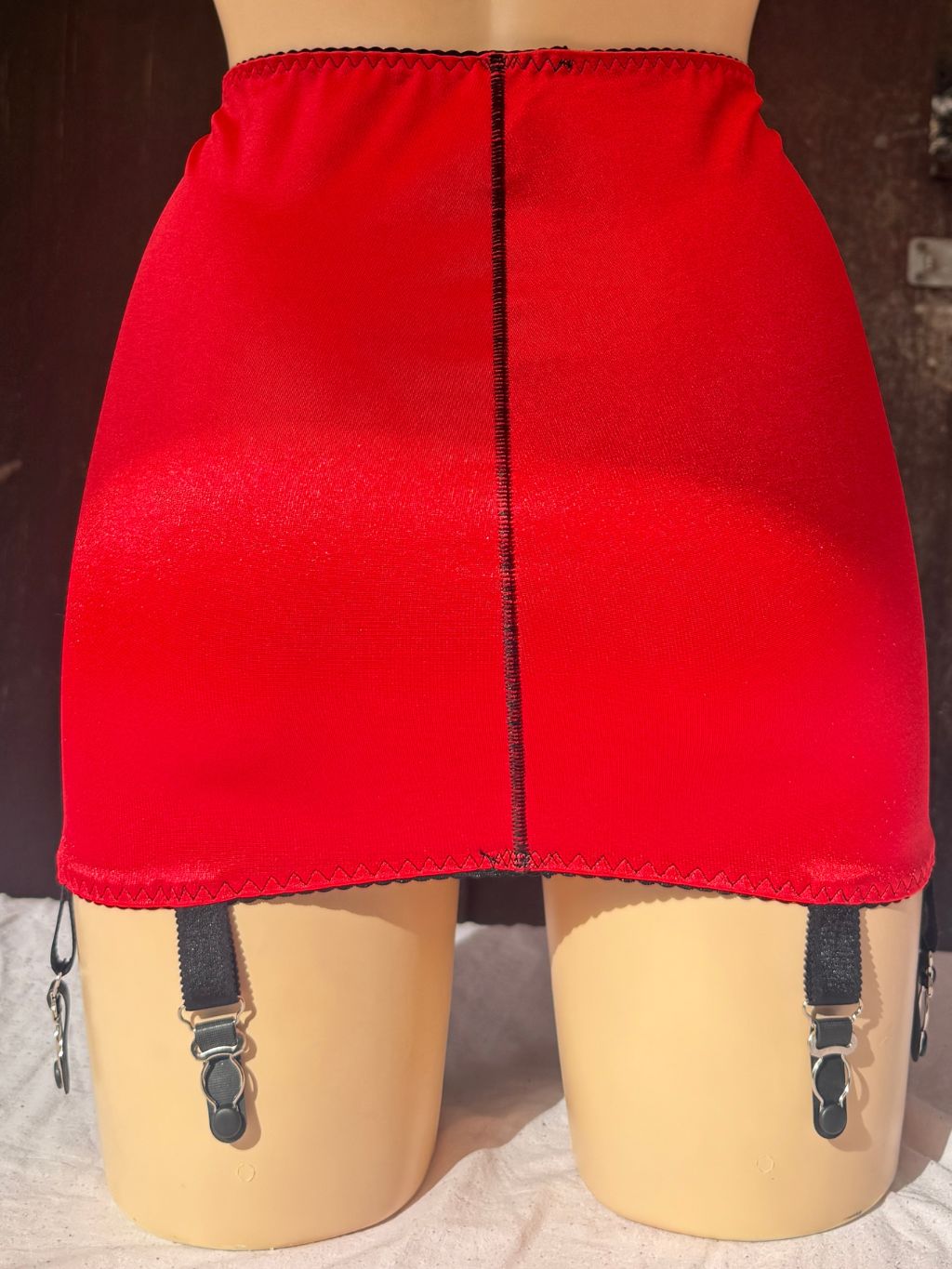 A bright red girdle displayed on a beige mannequin, featuring a central black seam and black suspender straps with silver metal clips hanging from the hem.