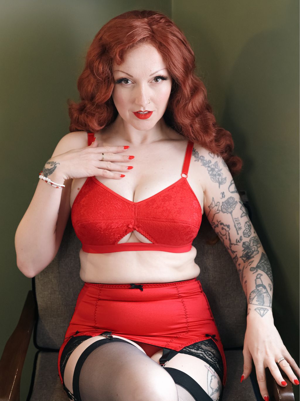 A woman with red hair and tattoos wearing a red bra and matching red skirt, seated against a dark olive-green wall.