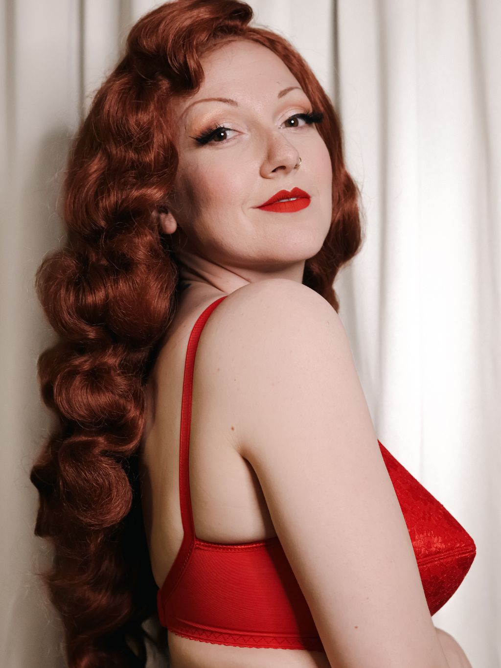 A woman with long, wavy red hair poses in a red bralette against a soft white curtain background.