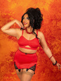 A woman modeling a red lingerie set consisting of a cutout bralette and a matching high-waisted garter skirt with black lace trim, posed against a textured orange and yellow background.