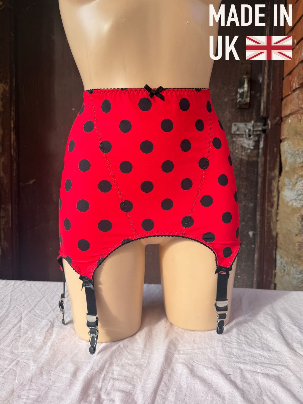 Red  roll on girdle with black polka dots and attached black garter straps, displayed on a beige mannequin torso. A 'MADE IN UK' graphic overlay is in the top right corner.