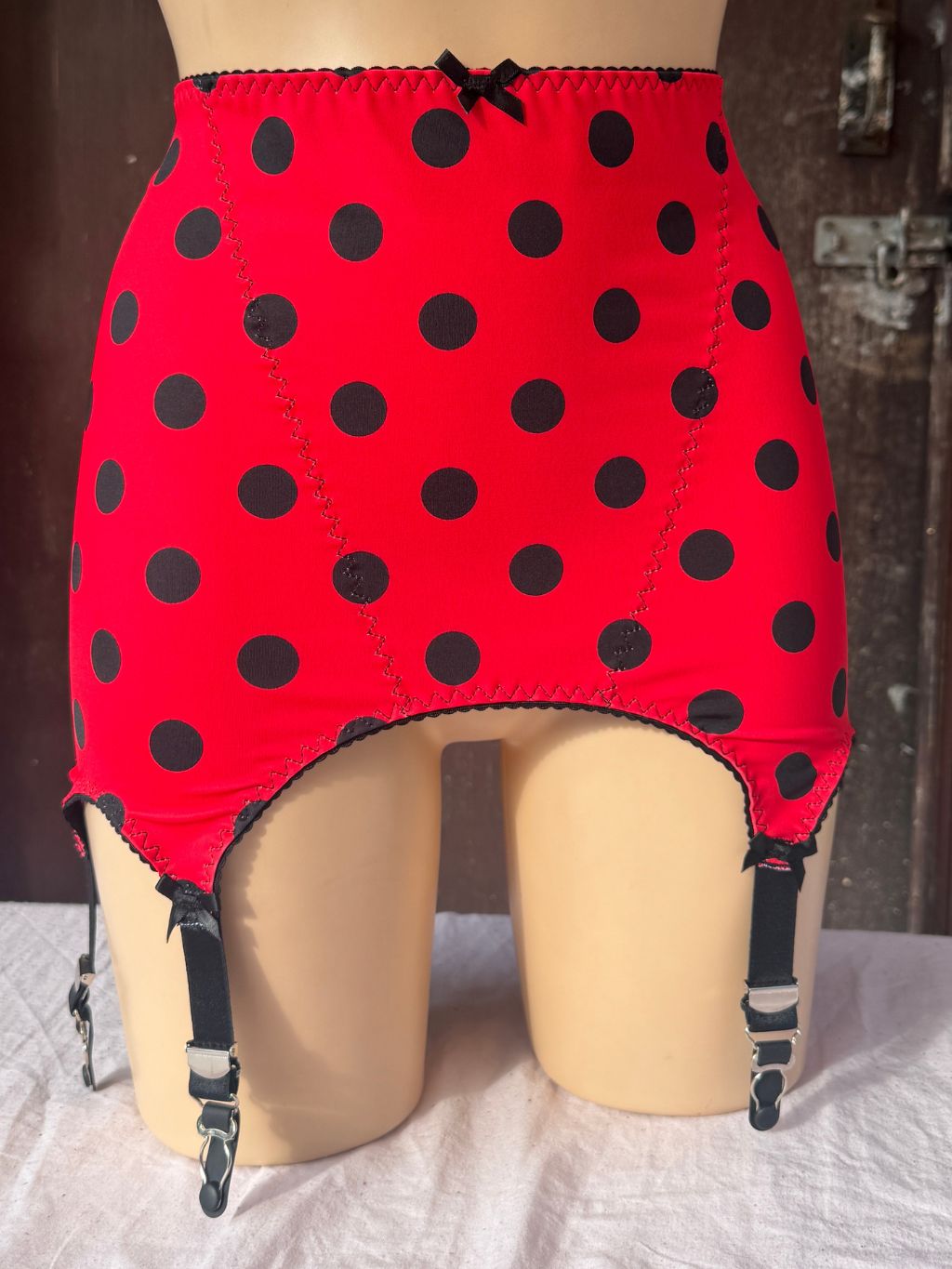 Red roll on girdle with black polka dots and attached black garter straps, displayed on a beige mannequin against a rustic dark wood background.