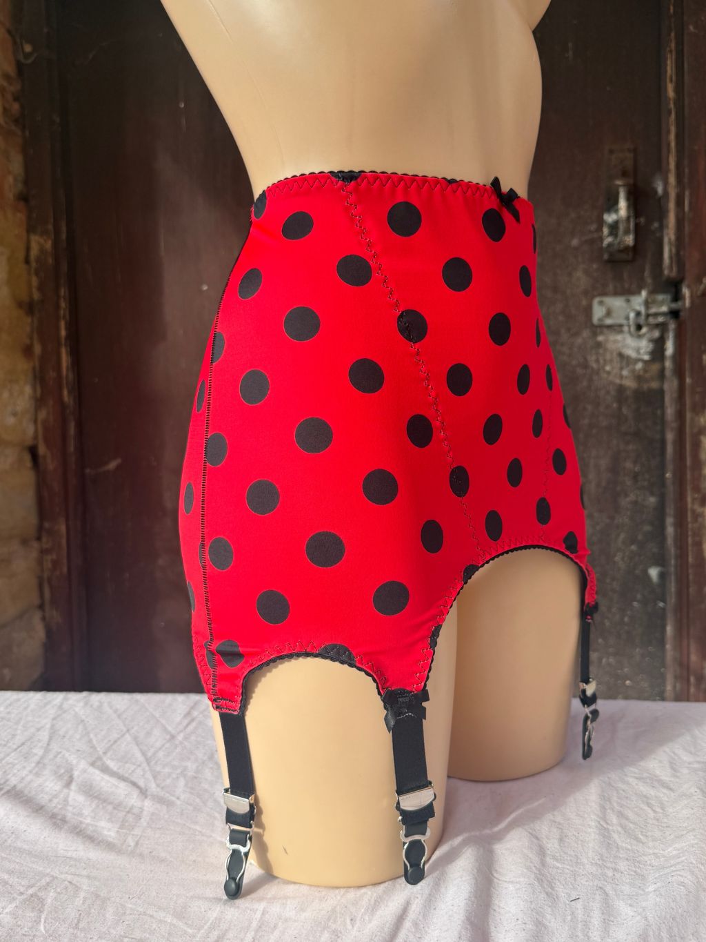 Red  roll on girdle  with black polka dots and attached black garter straps, displayed on a beige mannequin torso against a rustic dark wood door background.