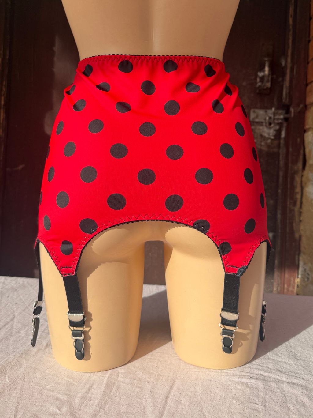 Red  roll on girdle  with black polka dots and attached black garter straps, displayed on a beige mannequin torso against a rustic wooden door background.
