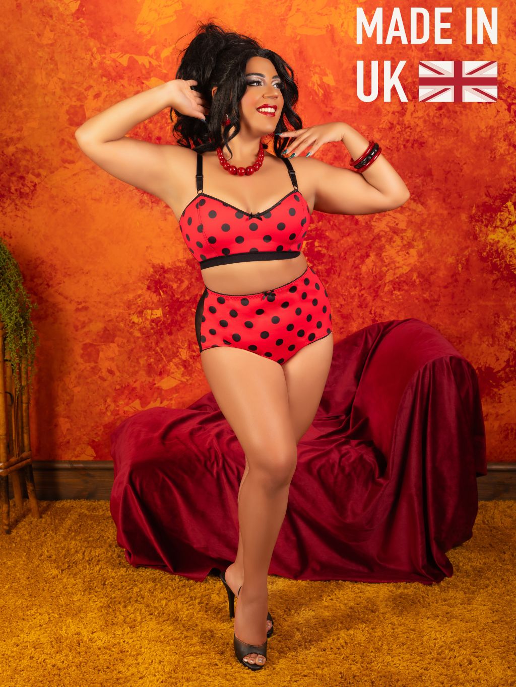 A woman models a red and black polka-dot lingerie set consisting of a bralette and matching high-waisted briefs, posed in front of a textured orange wall with a draped red velvet sofa. The text 'MADE IN UK' with a Union Jack flag is visible in the top right corner.