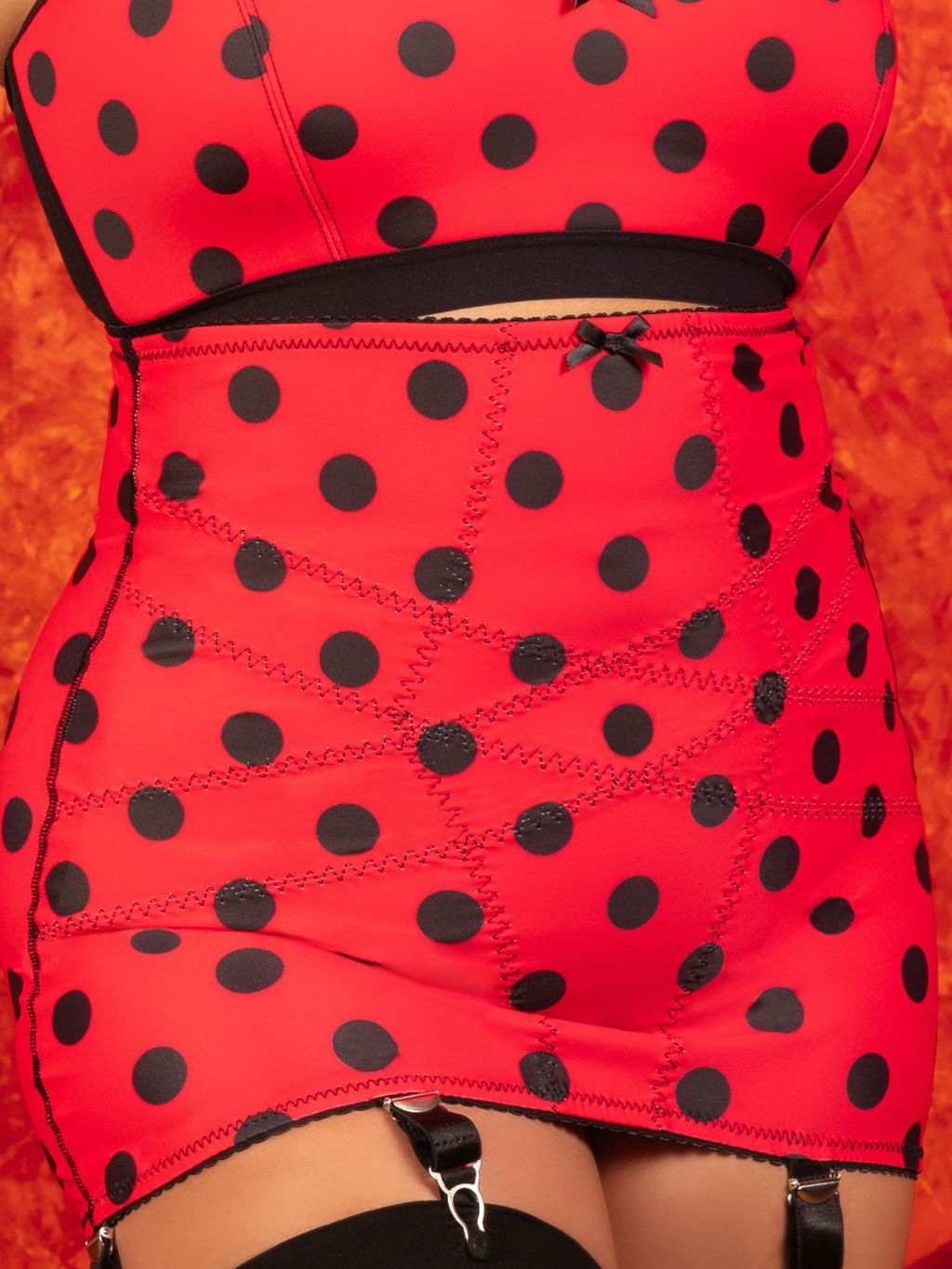 Red and black polka dot  bra and girdle, shown on a model against a textured orange background.