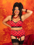 A woman posing in a red and black polka dot lingerie set with matching garter belt and thigh-high stockings, set against a textured orange and red background.