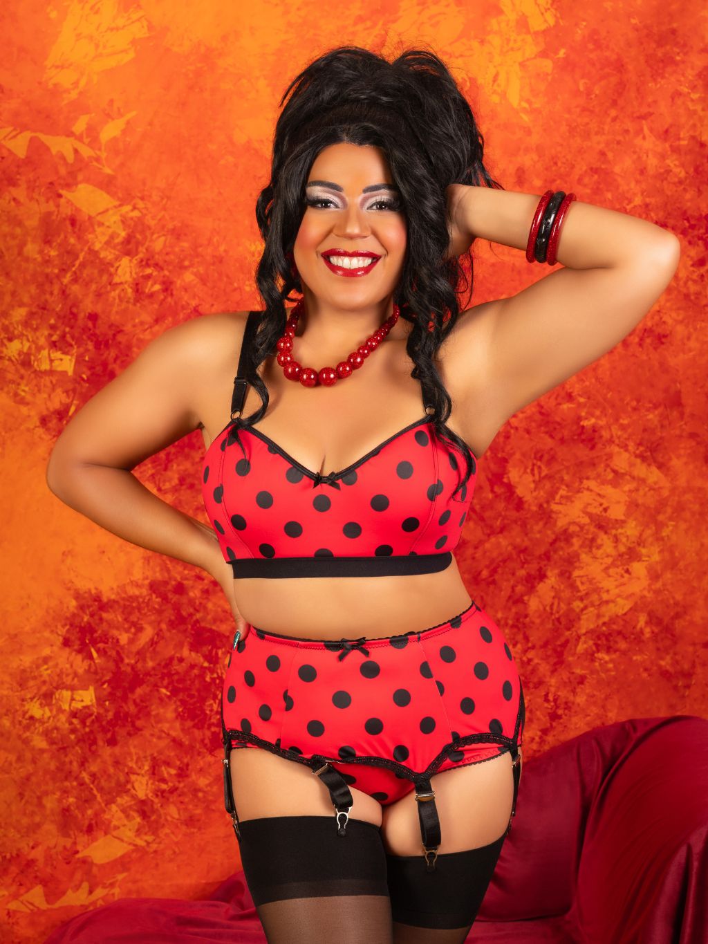 A woman posing in a red and black polka dot lingerie set with matching garter belt and thigh-high stockings, set against a textured orange and red background.