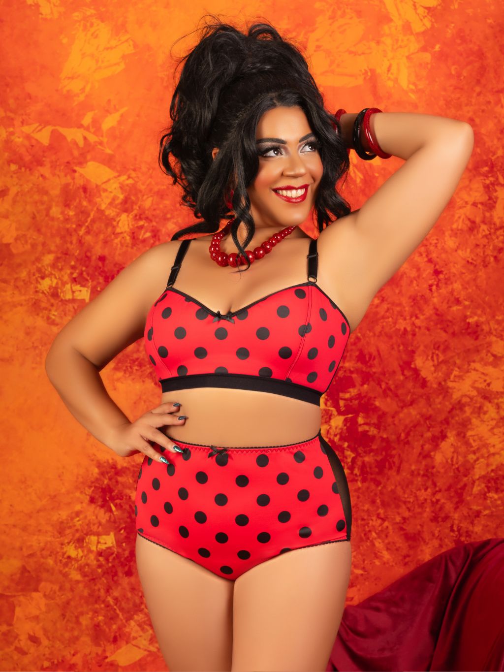 A woman modeling a red and black polka dot two-piece lingerie set against a textured orange background.