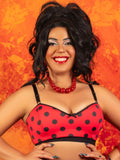 A smiling woman with dark curly hair poses in a red and black polka-dot bra top against a textured orange background.