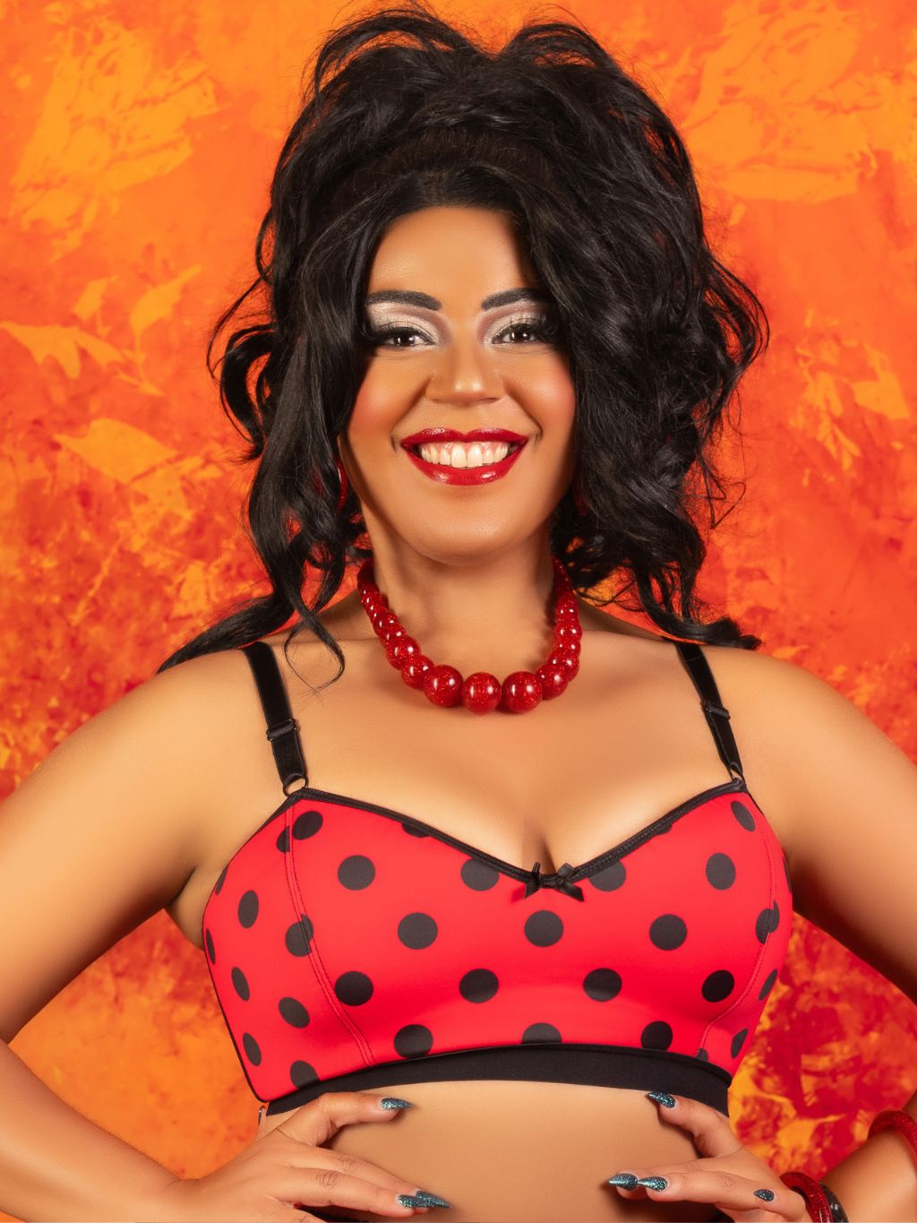 A smiling woman with dark curly hair poses in a red and black polka-dot bra top against a textured orange background.