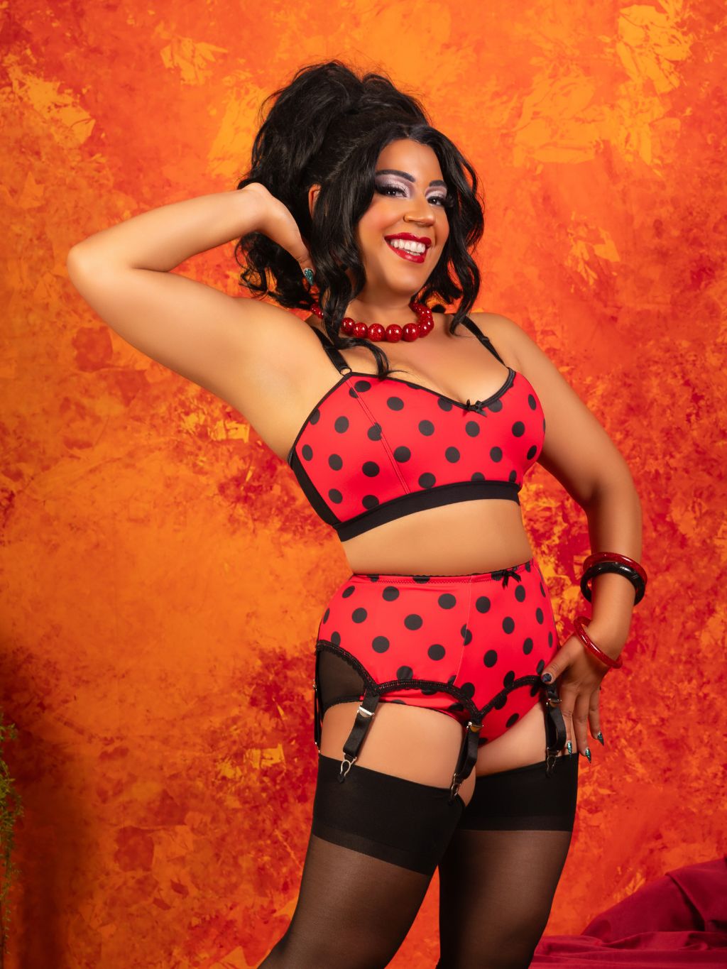 A woman modeling a red and black polka dot lingerie set with garter straps and black thigh-high stockings, posing against a textured orange background.