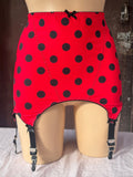 Red roll on girdle with black polka dots and black garter straps, displayed on a beige mannequin torso against a rustic wooden background.
