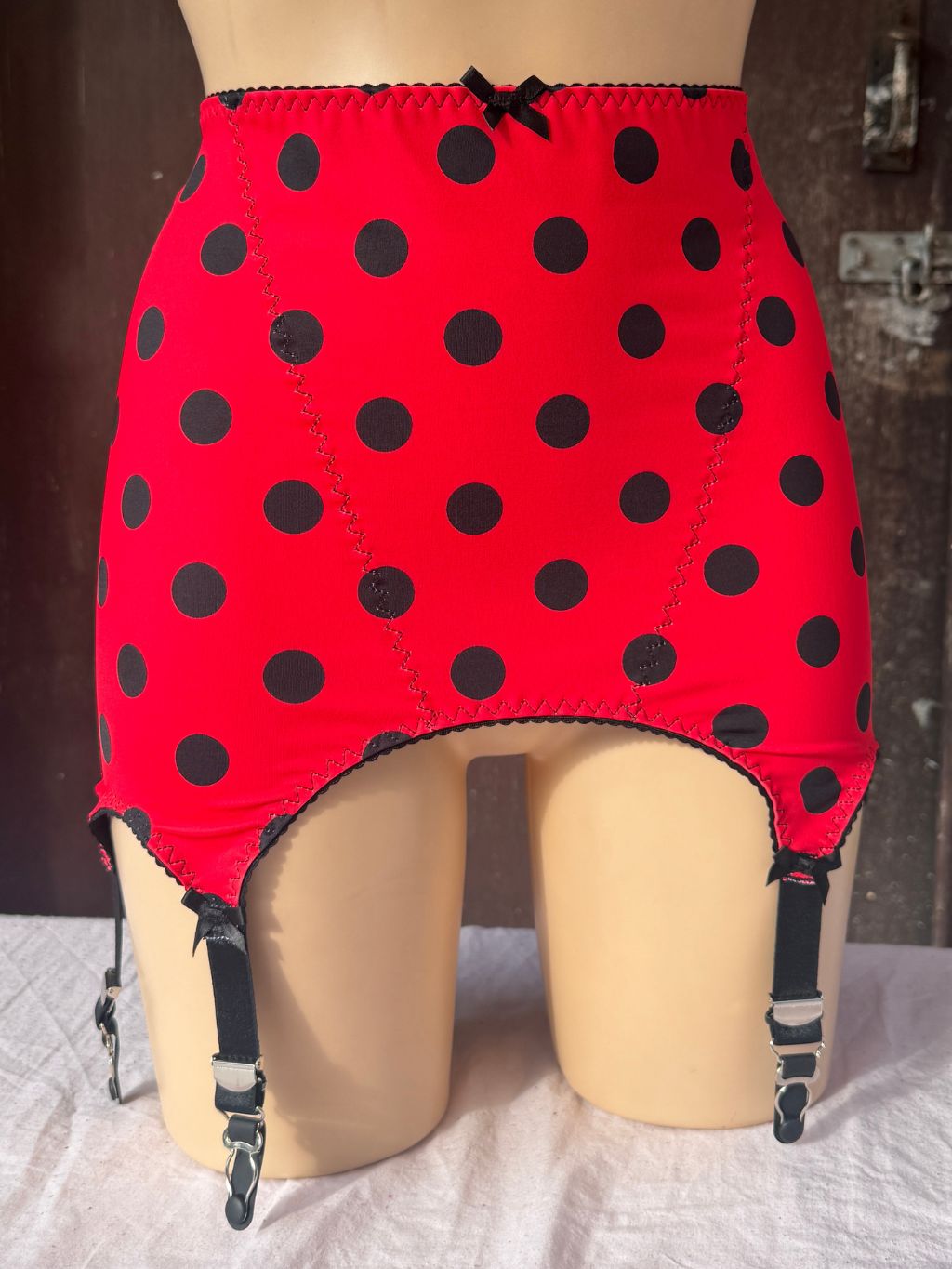 Red roll on girdle with black polka dots and black garter straps, displayed on a beige mannequin torso against a rustic wooden background.