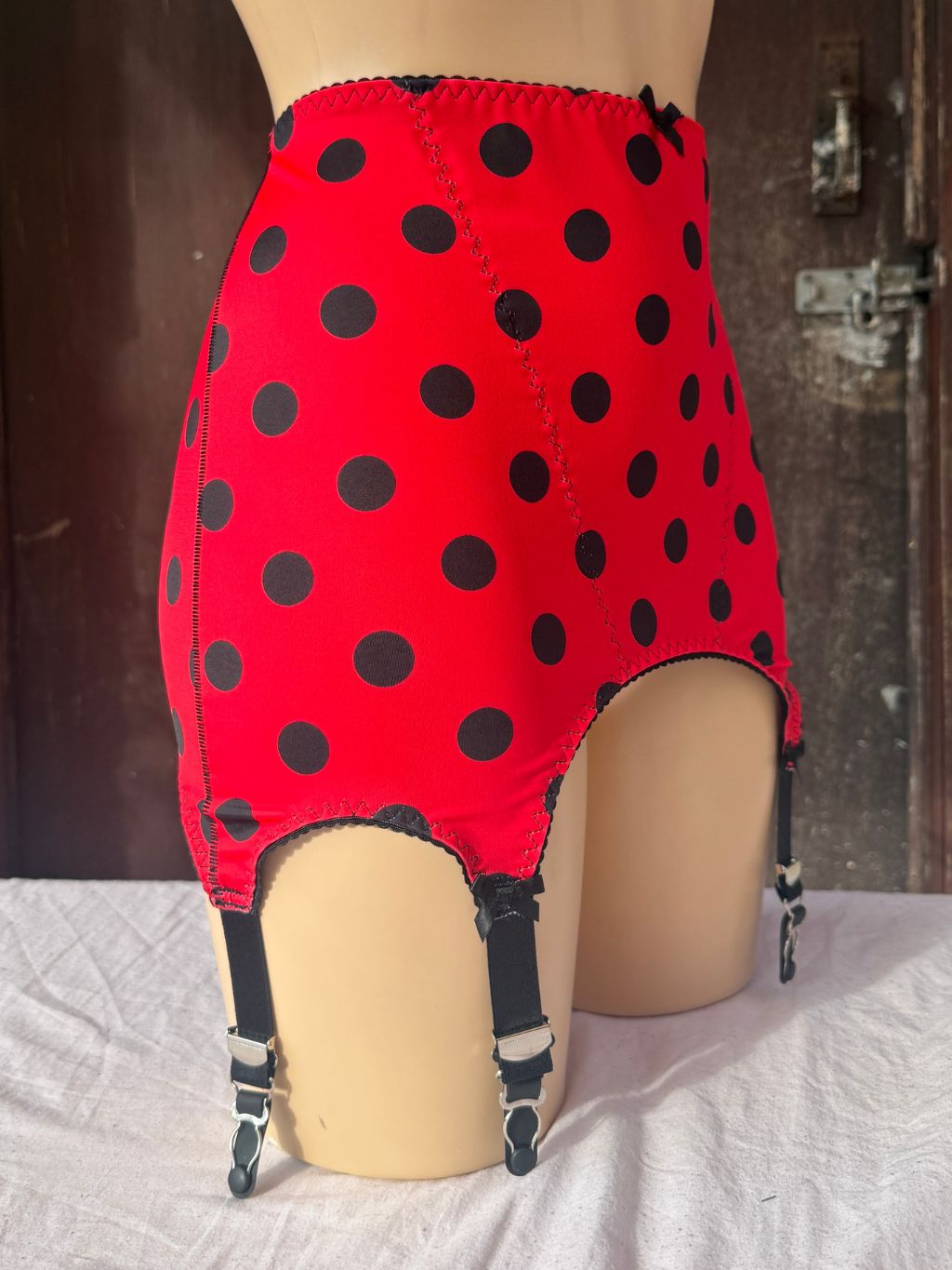Red roll on girdle with black polka dots and attached black garter straps, displayed on a beige mannequin against a rustic dark wood door background.