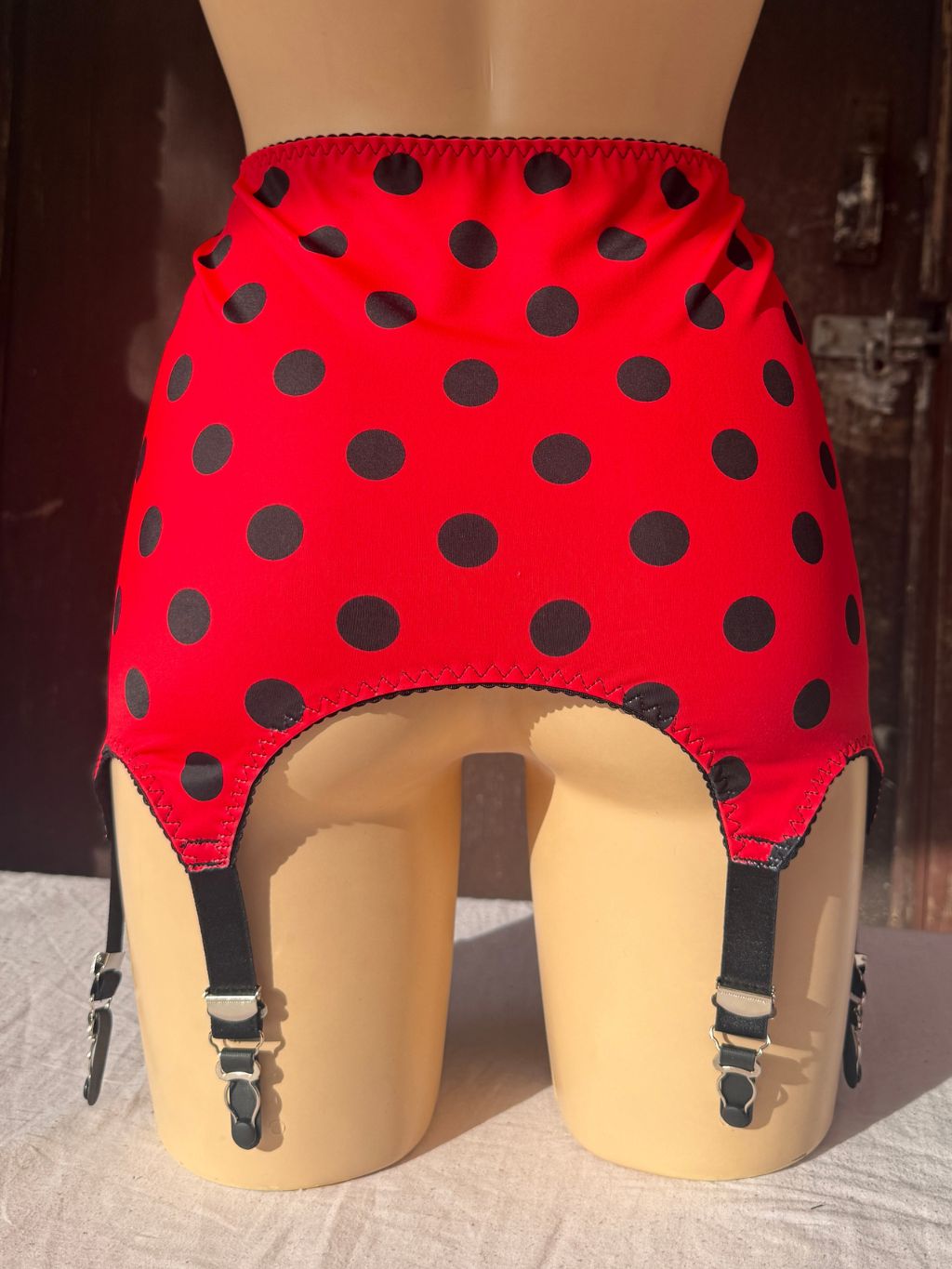 Red girdle with black polka dots and attached black garter straps, displayed on a beige mannequin lower body against a dark wooden background.