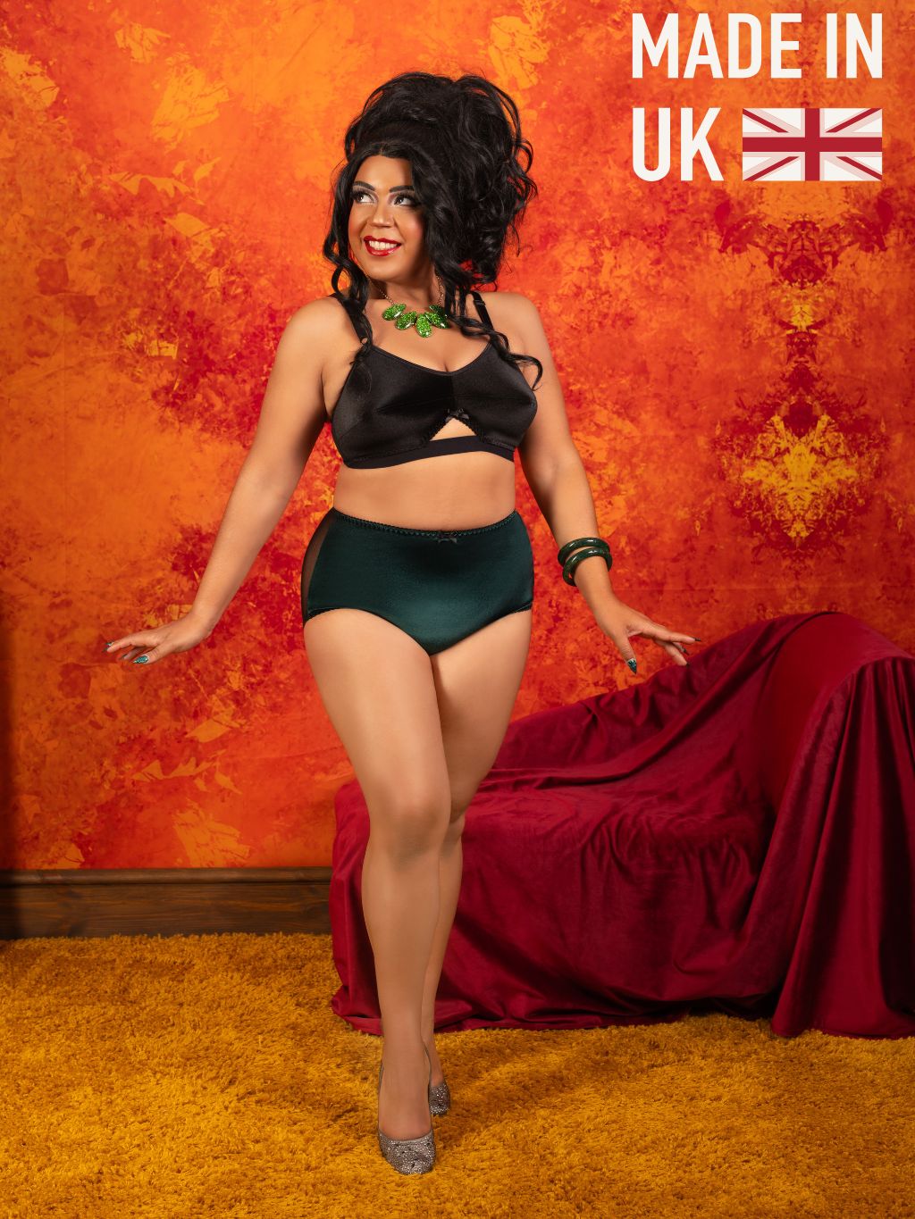 A woman models a black satin halter bra and matching dark green satin briefs against a textured orange and yellow studio backdrop with a draped red sofa and a 'MADE IN UK' graphic in the top right corner.