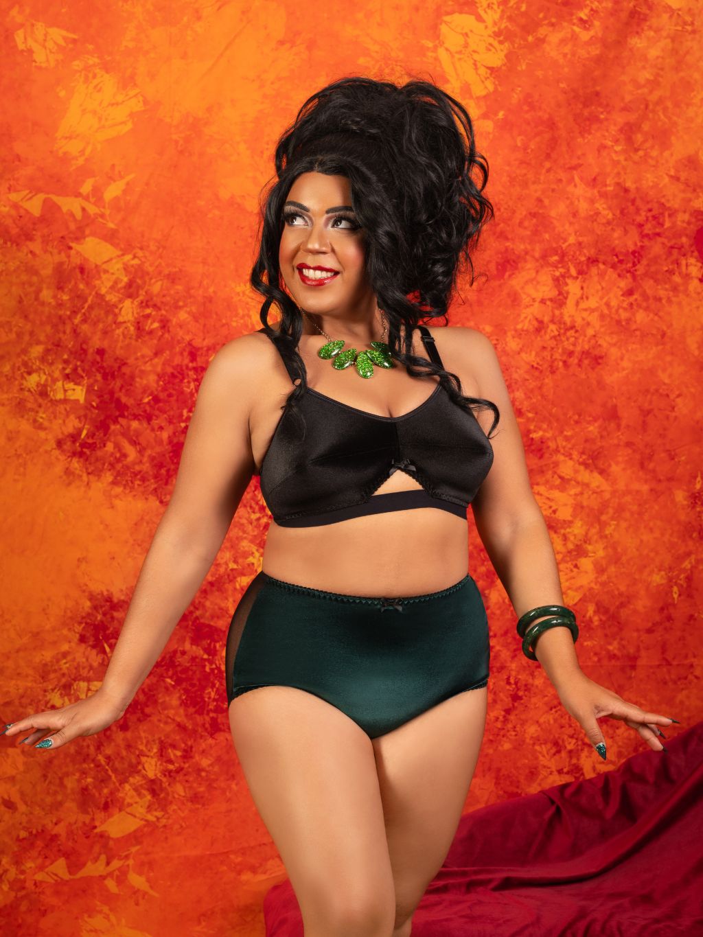A woman modeling a black satin bralette and dark green high-waisted briefs against a textured orange and red background.