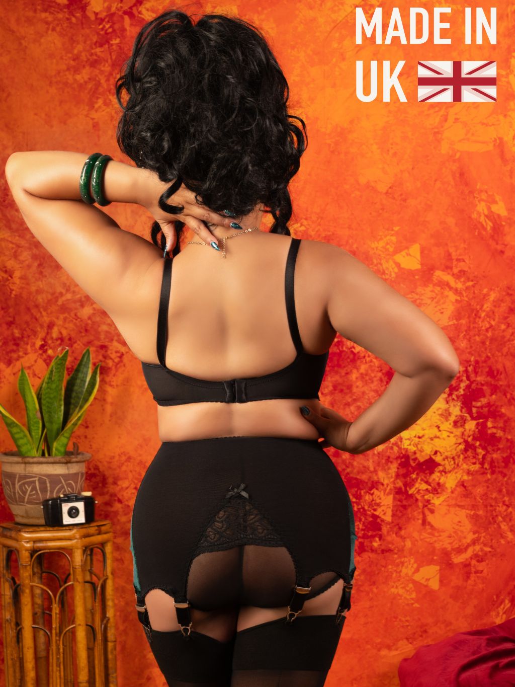 A woman shown from the back wearing black lingerie, including a bra, high-waisted briefs with lace and garter details, and thigh-high stockings, posed against a textured orange and red wall. A small plant and side table are visible on the left, and 'MADE IN UK' with a Union Jack appears in the top right corner.