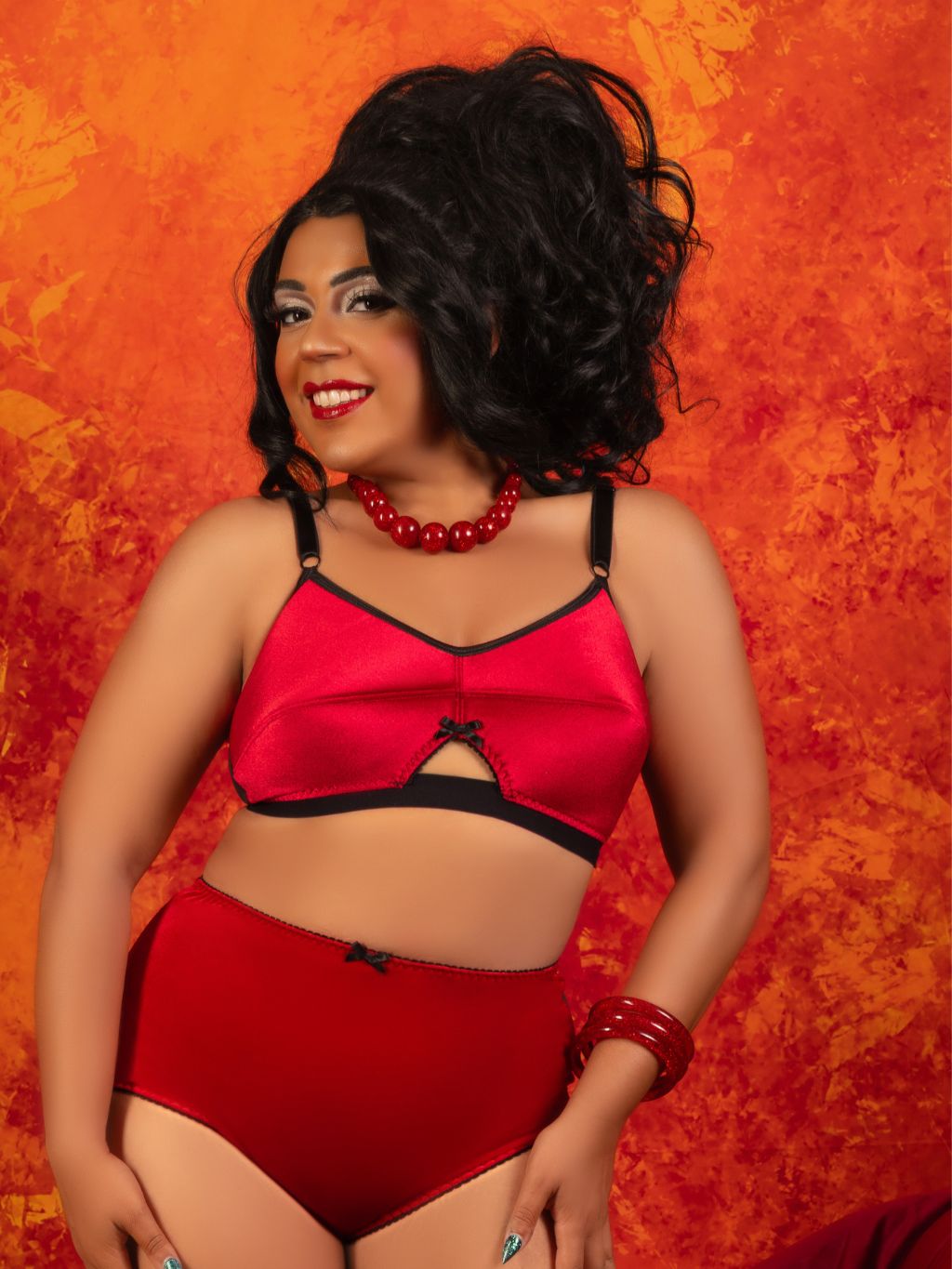A woman modeling a red and black lingerie set against a textured orange and red background. The set includes a satin-like red bra top with black trim and matching high-waisted red briefs.